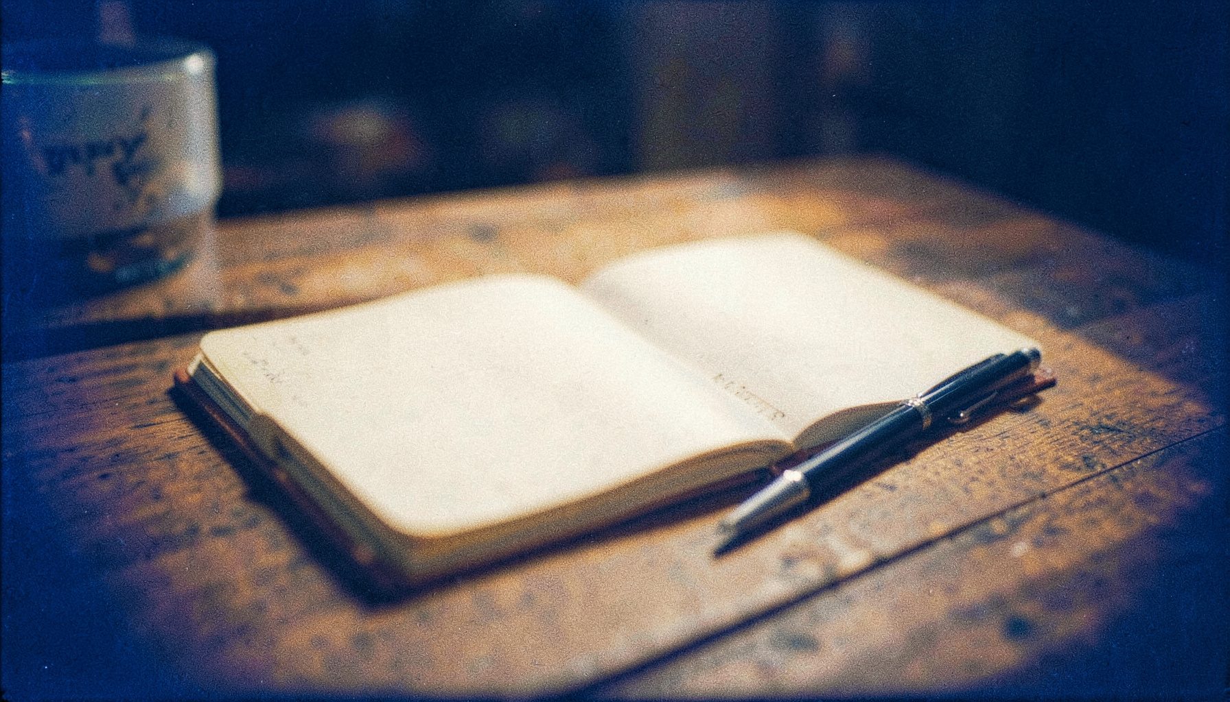 A grainy analog film photograph of a warm, softly lit wooden tabletop with an open notebook and a pen, bathed in gentle golden-hour light with deep navy shadows at the edges.