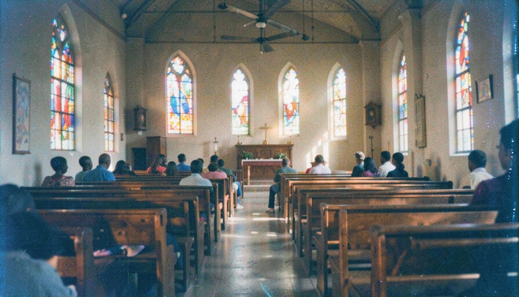 An analog film photograph with visible grain depicting a warm-toned African church interior with colorful stained glass and soft natural light spilling across rows of wooden benches, conveying community and devotion.