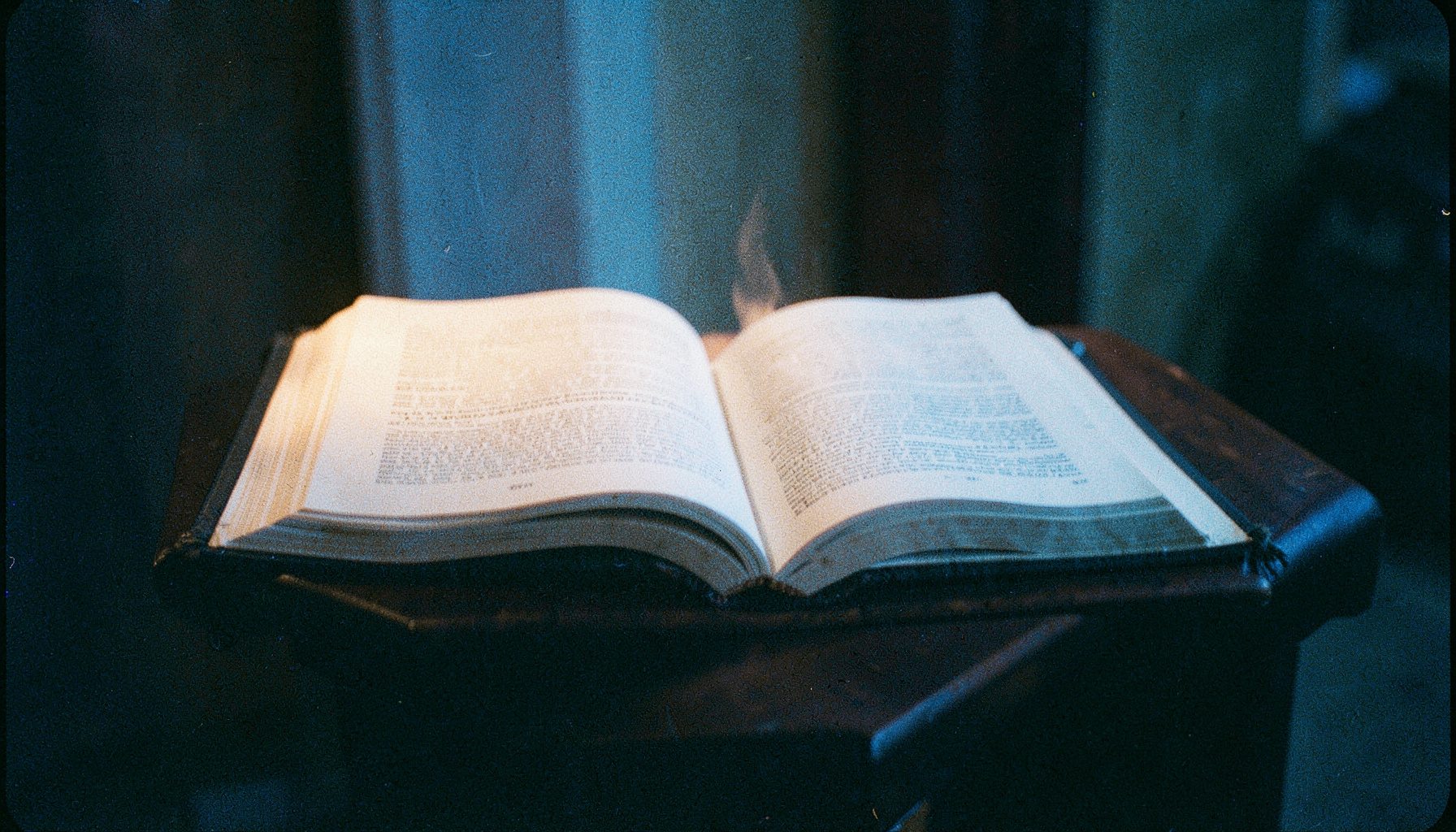 An analog film photograph with visible grain of an open Bible on a wooden lectern, soft candlelight casting gentle shadows, evoking quiet study and spiritual reflection.