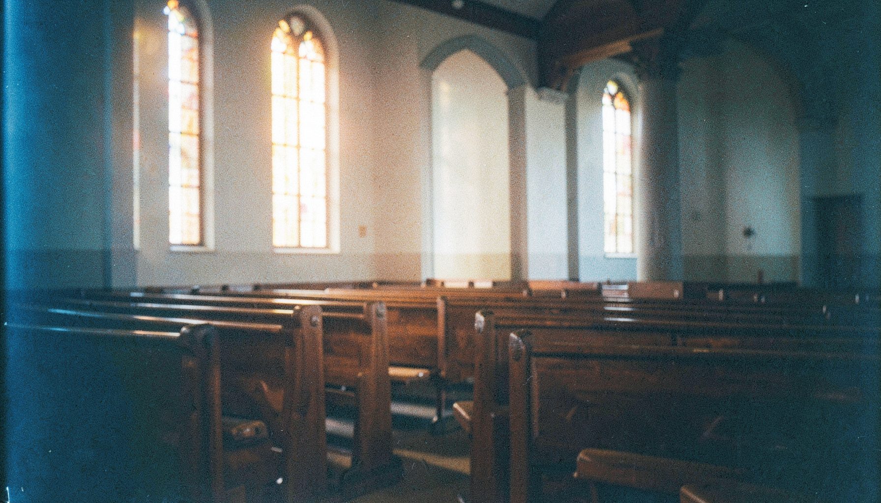 A warm analog film photograph with visible grain showing a softly lit worship space with wooden pews and gentle amber light filtering through tall windows, creating a reverent and peaceful atmosphere.
