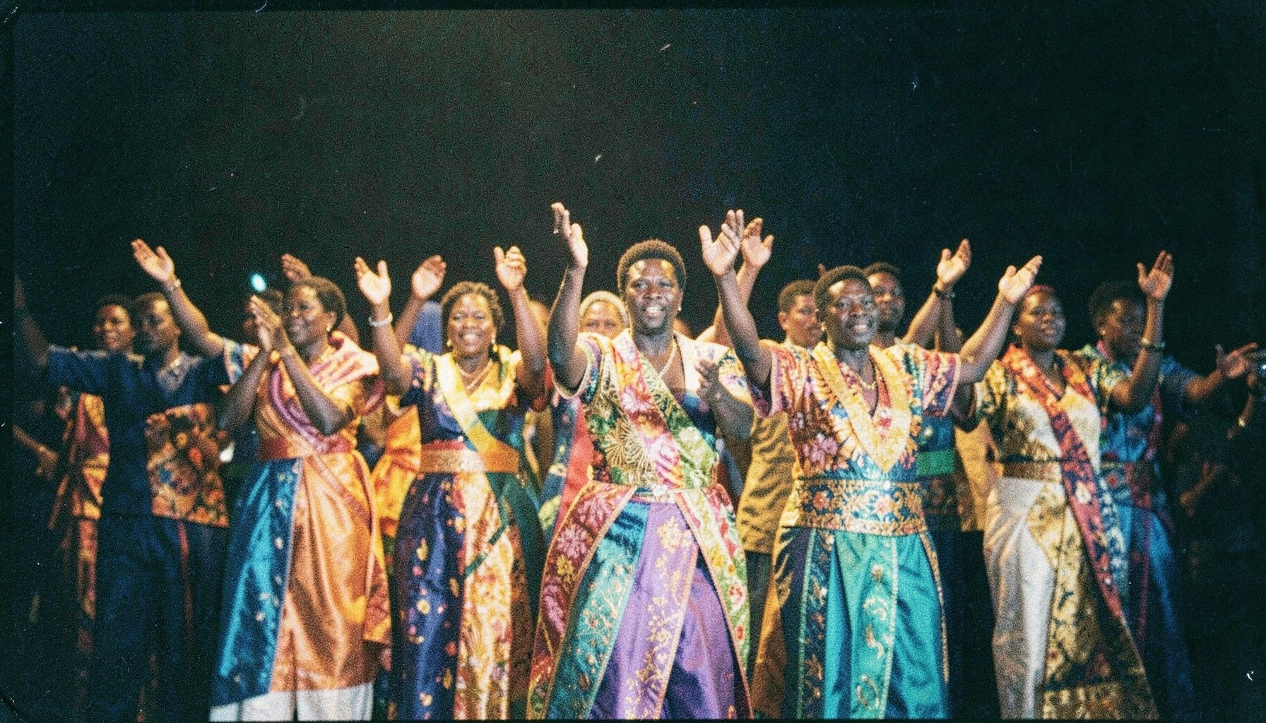 An analog film photograph with visible grain showing a Nigerian worship choir in colorful traditional attire, hands raised, bathed in warm golden stage light against a dark background.