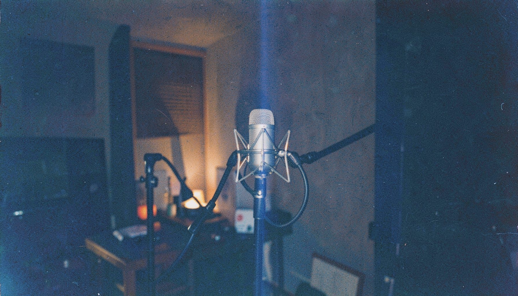 A warm, grainy film photograph of a dimly lit recording studio with a microphone on a stand, soft amber and navy shadows, moody and intimate atmosphere.