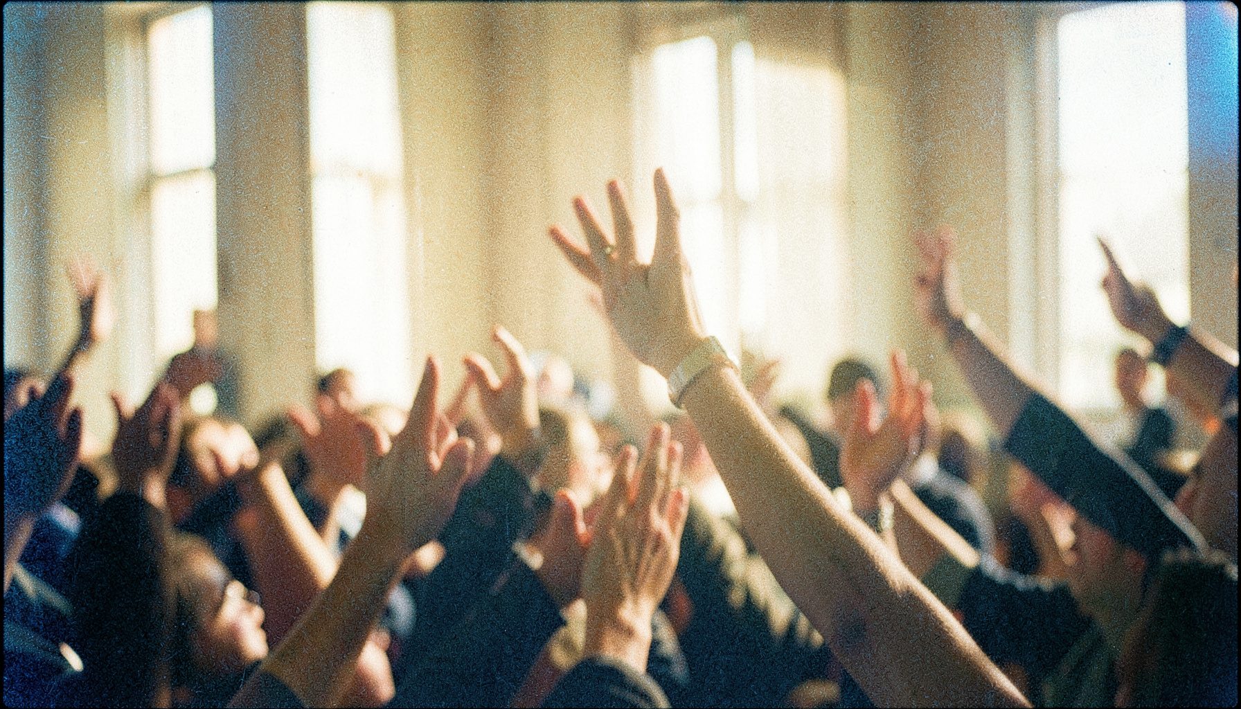 A grainy analog film photograph of hands lifted in a congregation, soft natural light streaming through high windows, warm ochre and cream tones