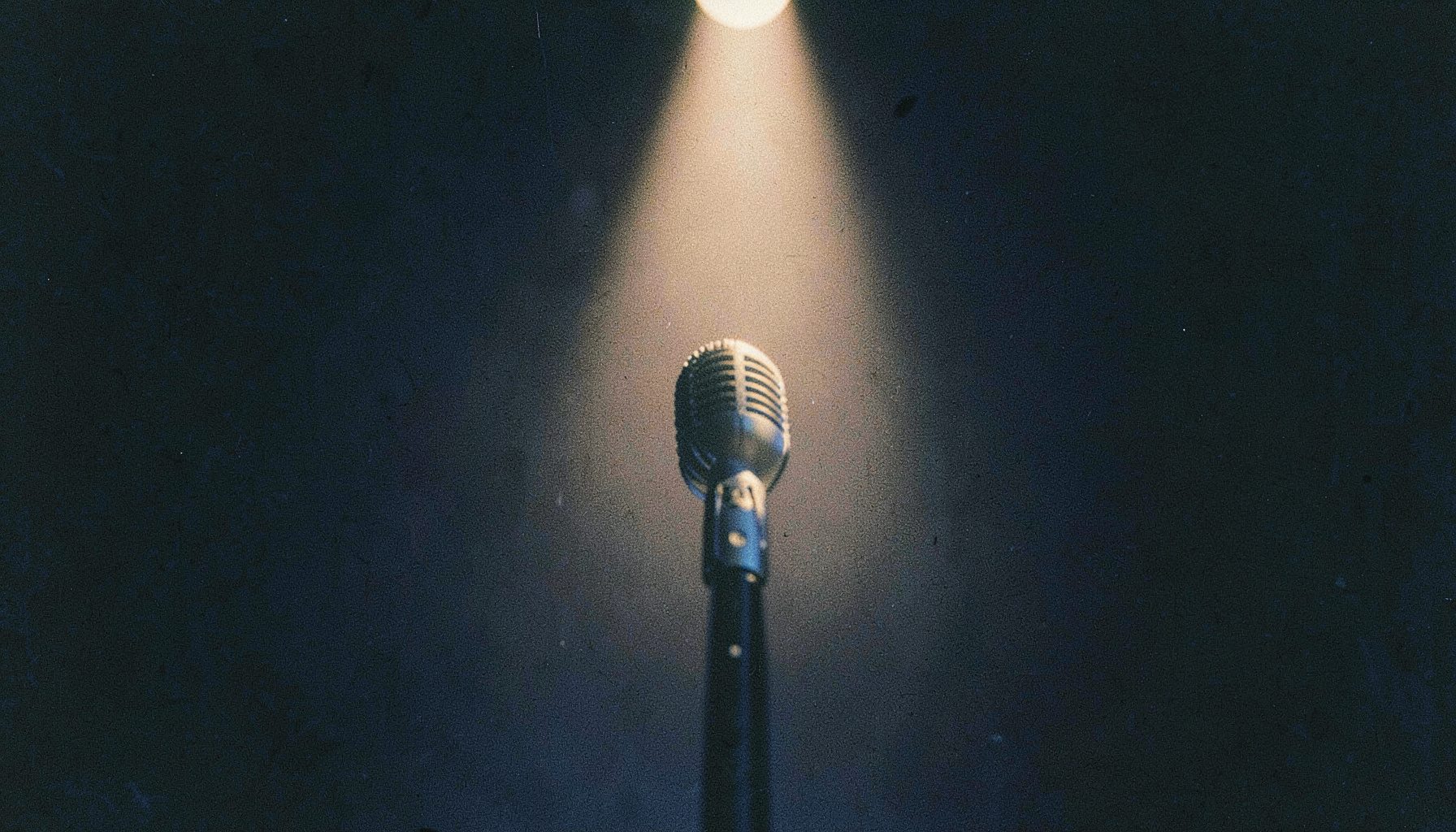 A grainy analog film photograph of a microphone on a dark stage with a single warm spotlight, deep blacks and soft cream tones