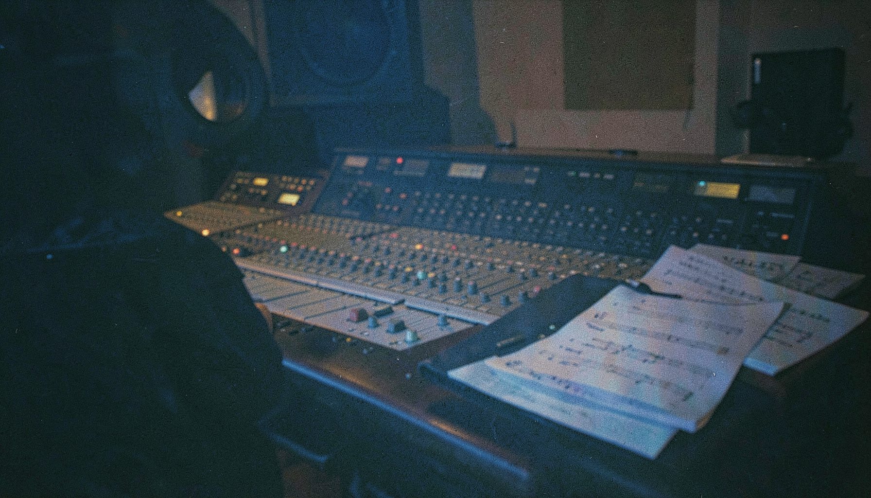A grainy analog film photograph of a recording studio console bathed in dim blue and amber light, with sheet music resting nearby, moody and contemplative
