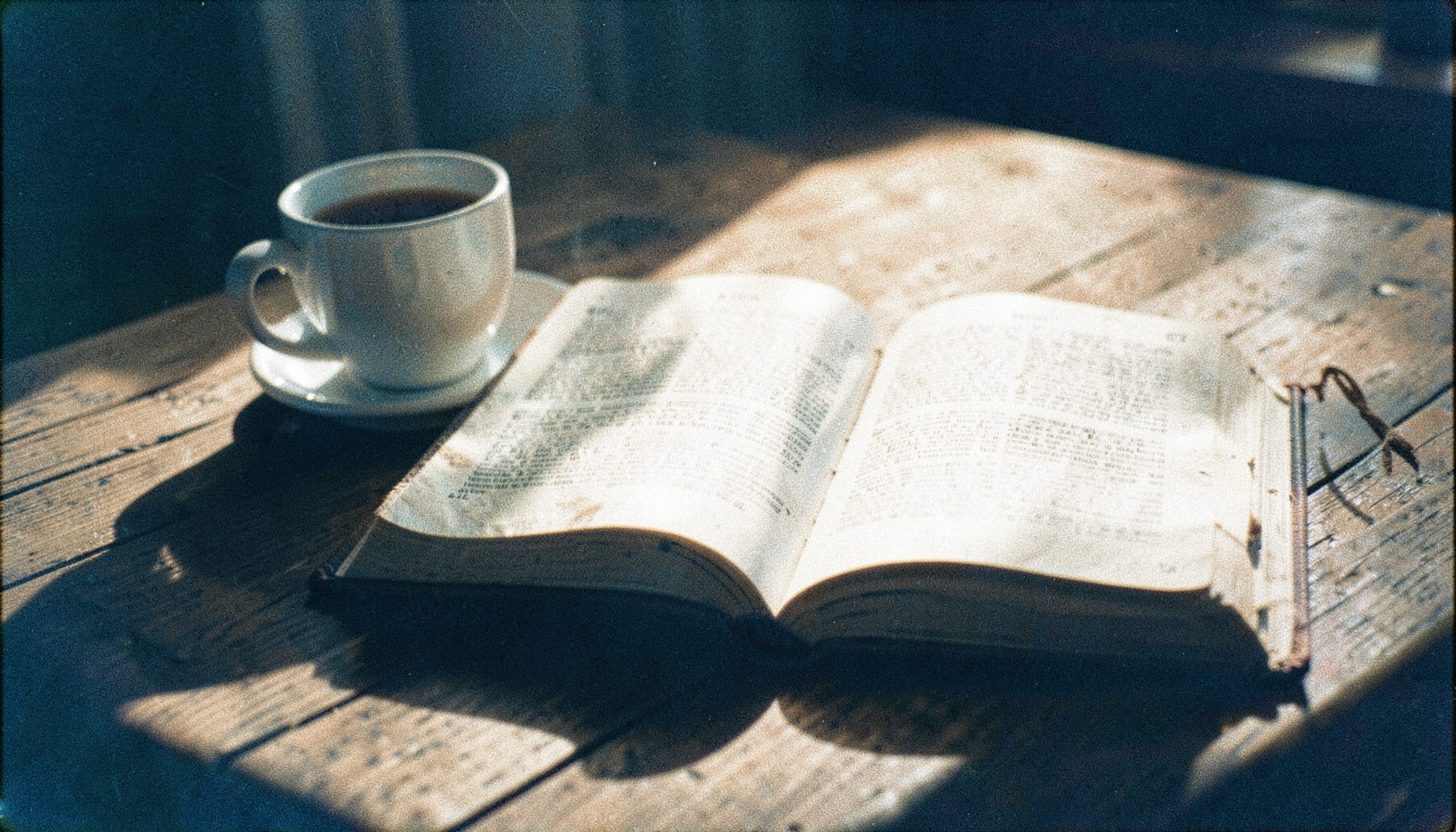 A grainy analog film photograph of a worn Bible open on a wooden table beside a cup of tea, morning light casting long shadows, sepia and cream tones