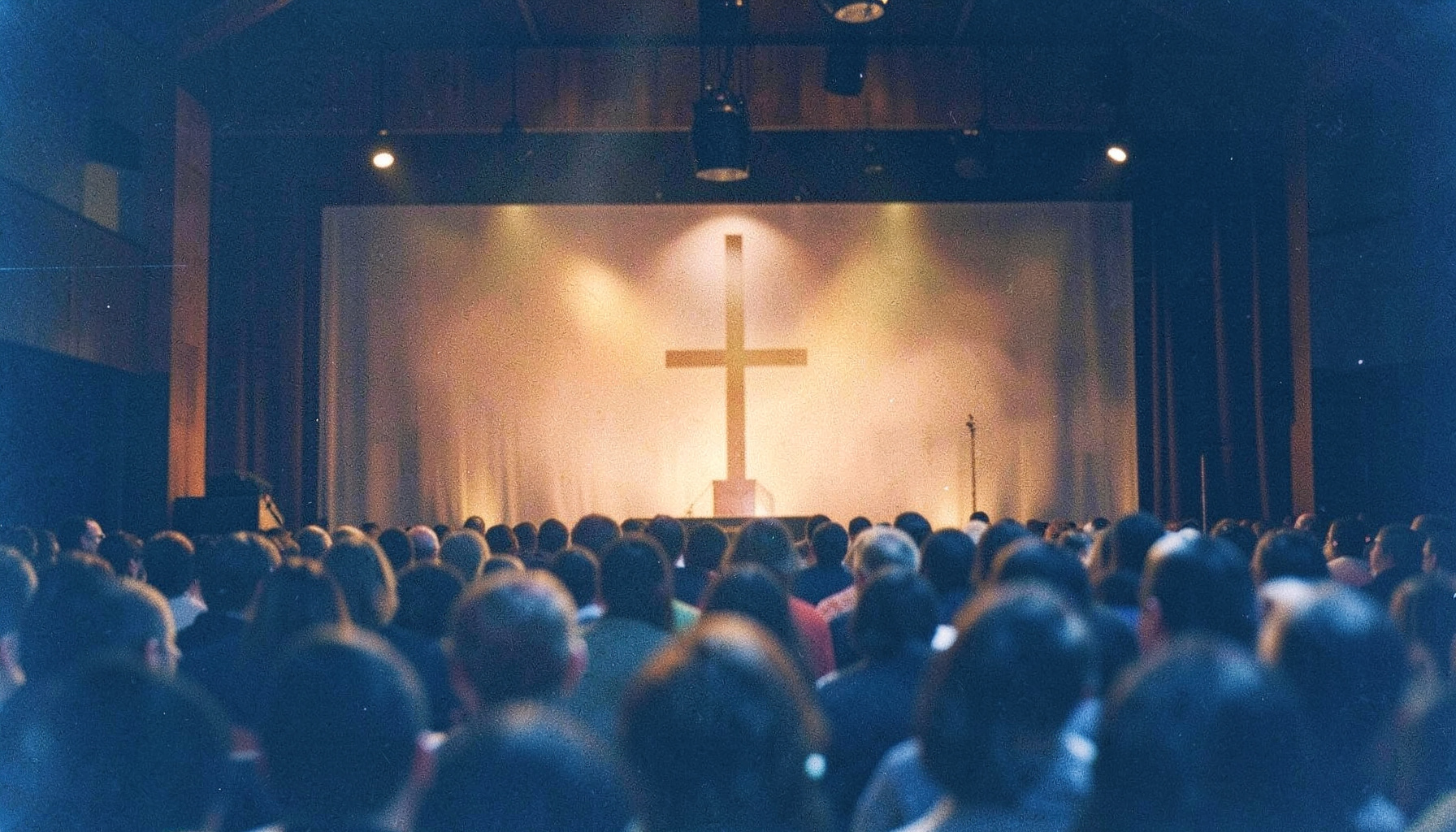 Analog film photograph of a worship conference stage with warm stage lighting and a cross silhouette, congregation blurred in the foreground, rich amber and navy tones