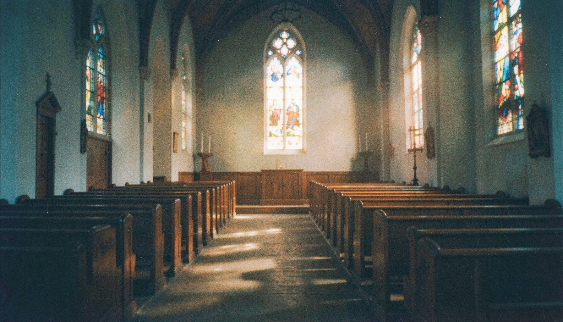 A warm analog film photograph of a softly lit church sanctuary with wooden pews and gentle amber light streaming through stained glass, quiet and reverent atmosphere with visible film grain