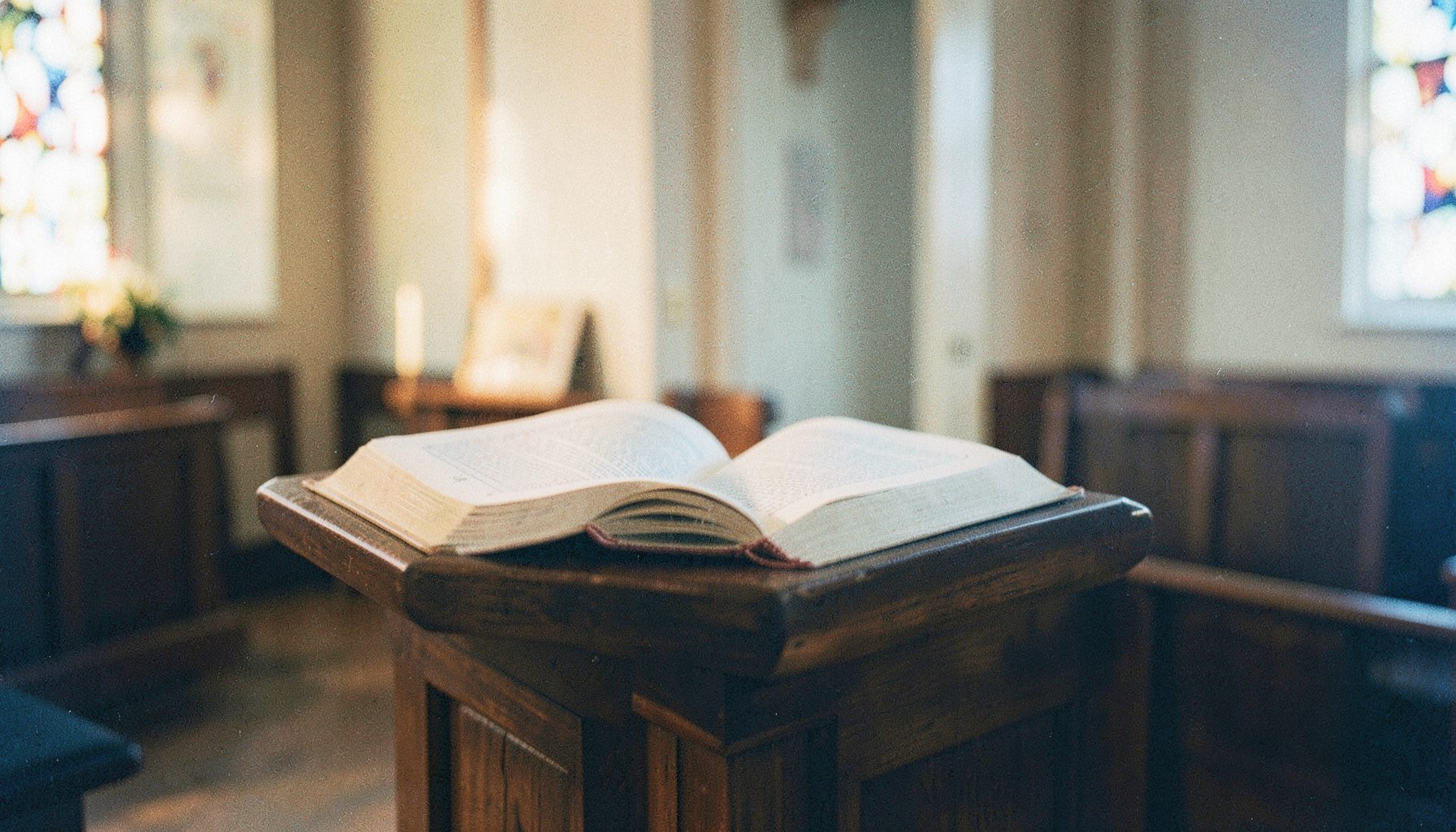A warm, softly lit analog film photograph of an open Bible resting on a wooden lectern in a quiet church interior, with gentle amber and cream tones and visible film grain.