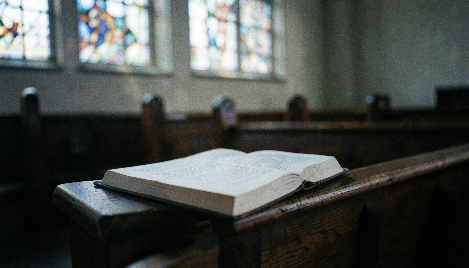 An open Bible resting on a wooden pew inside a quiet church, soft natural light streaming through stained glass, muted blues and ambers