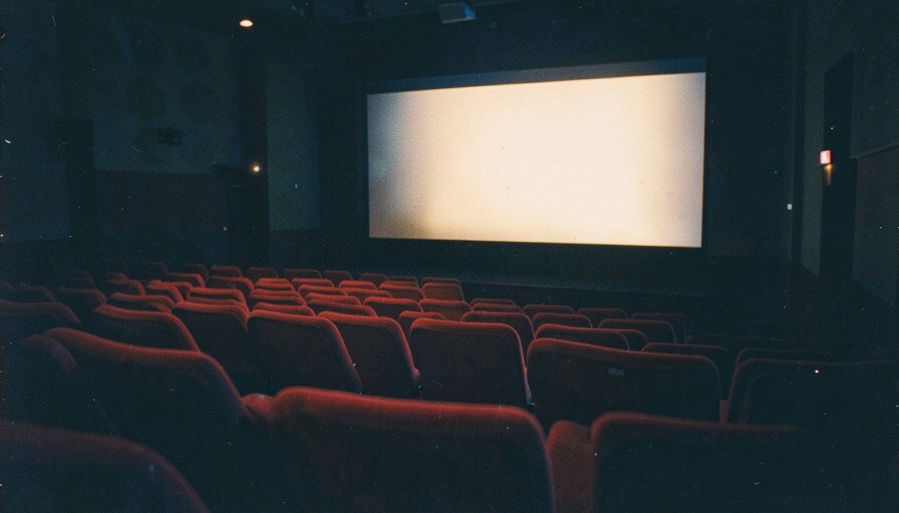 A dimly lit cinema auditorium with rows of red velvet seats, a glowing screen casting warm amber light across the empty room, analog film grain visible in the shadows