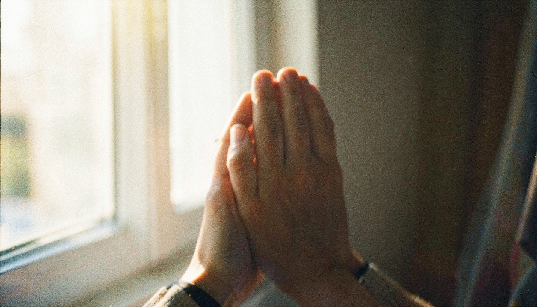 A soft-focus close-up of hands clasped in prayer against a sunlit window, warm golden tones with visible film grain