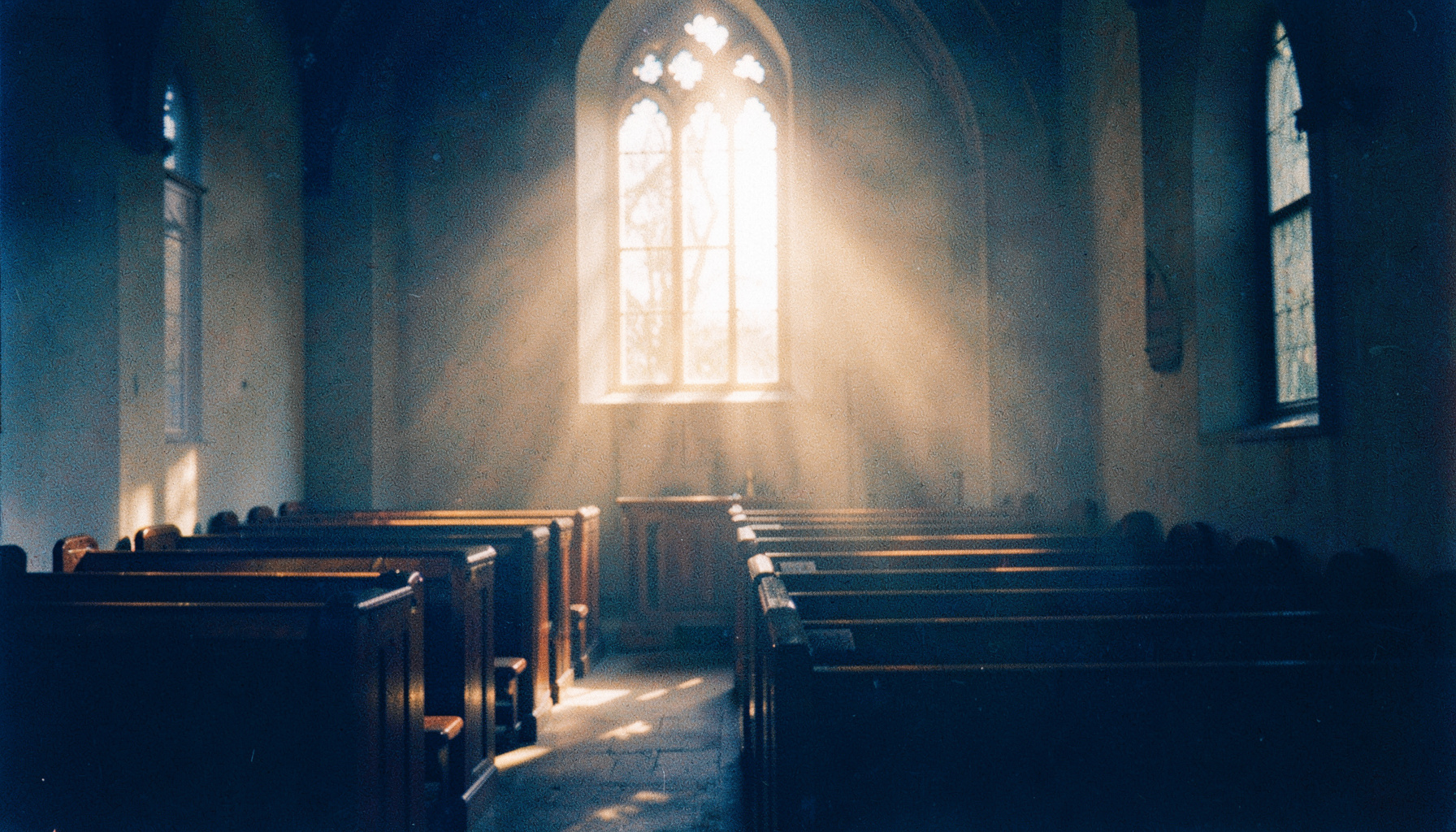 A warm, grainy film photograph of soft golden light streaming through a church window onto wooden pews, with a gentle haze and deep navy shadows.