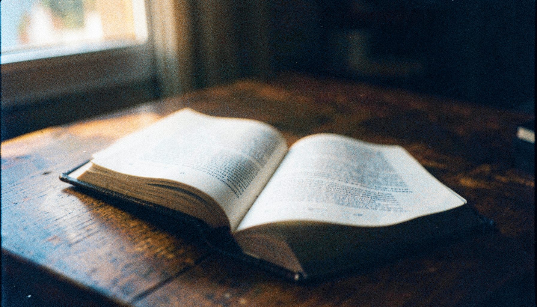 A grainy analog film photo of an open Bible resting on a wooden table, lit by soft window light with warm amber tones and deep brown shadows.