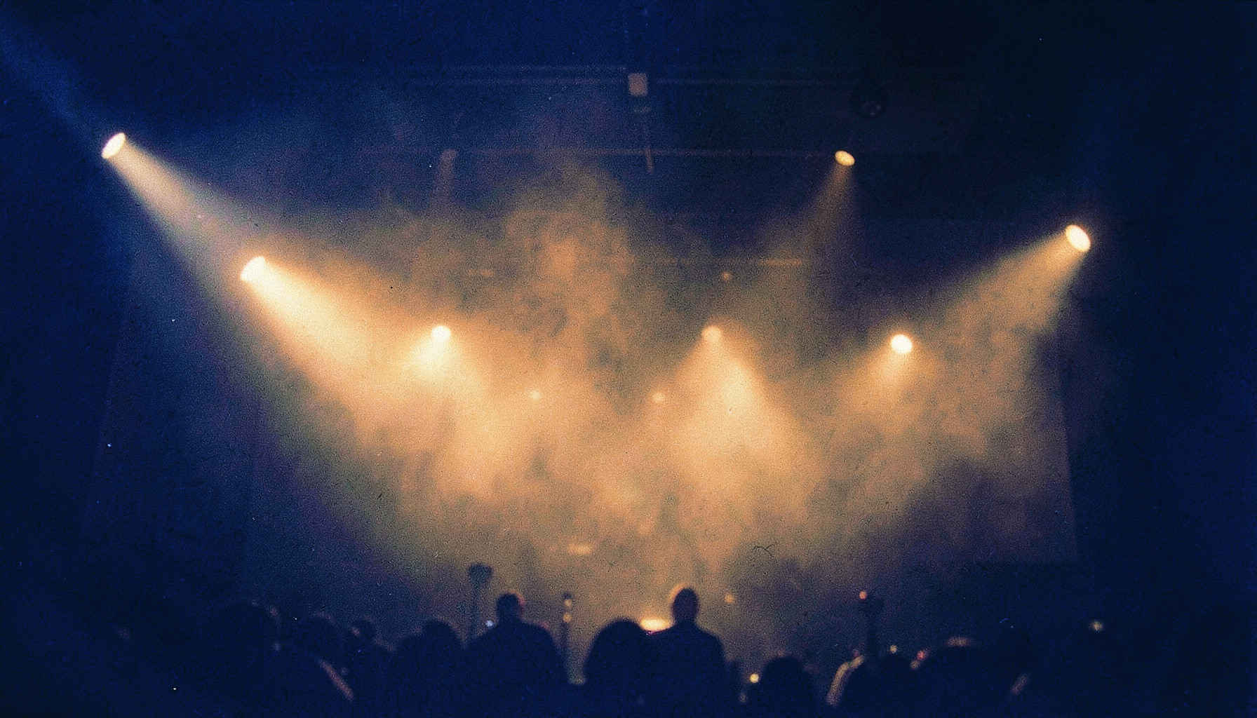 A grainy analog film photograph of warm amber stage lights glowing through atmospheric haze in a dim worship space, deep navy shadows and soft golden highlights
